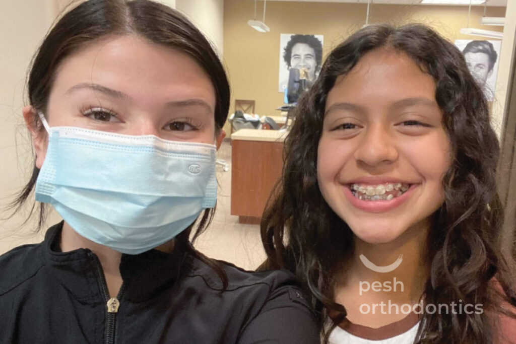 A teenage girl with a bright smile wearing new braces stands in the Pesh Orthodontics office in Menifee, California.