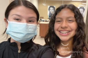 A teenage girl with a bright smile wearing new braces stands in the Pesh Orthodontics office in Menifee, California.